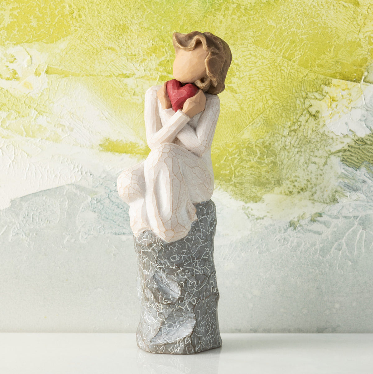 Willow Tree Always, 6”h hand-painted figure in cream dress, knees drawn up to chest, holding red heart to face, seated on gray rock etched with symbols of love, in front of lime painting