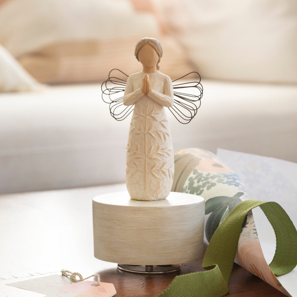 Willow Tree a tree, a prayer Musical, 7.5”h hand-painted resin angel figure in cream dress with wire wings, hands in prayer position, tree carving on dress, atop round, cream base containing musical element, on table with ribbon