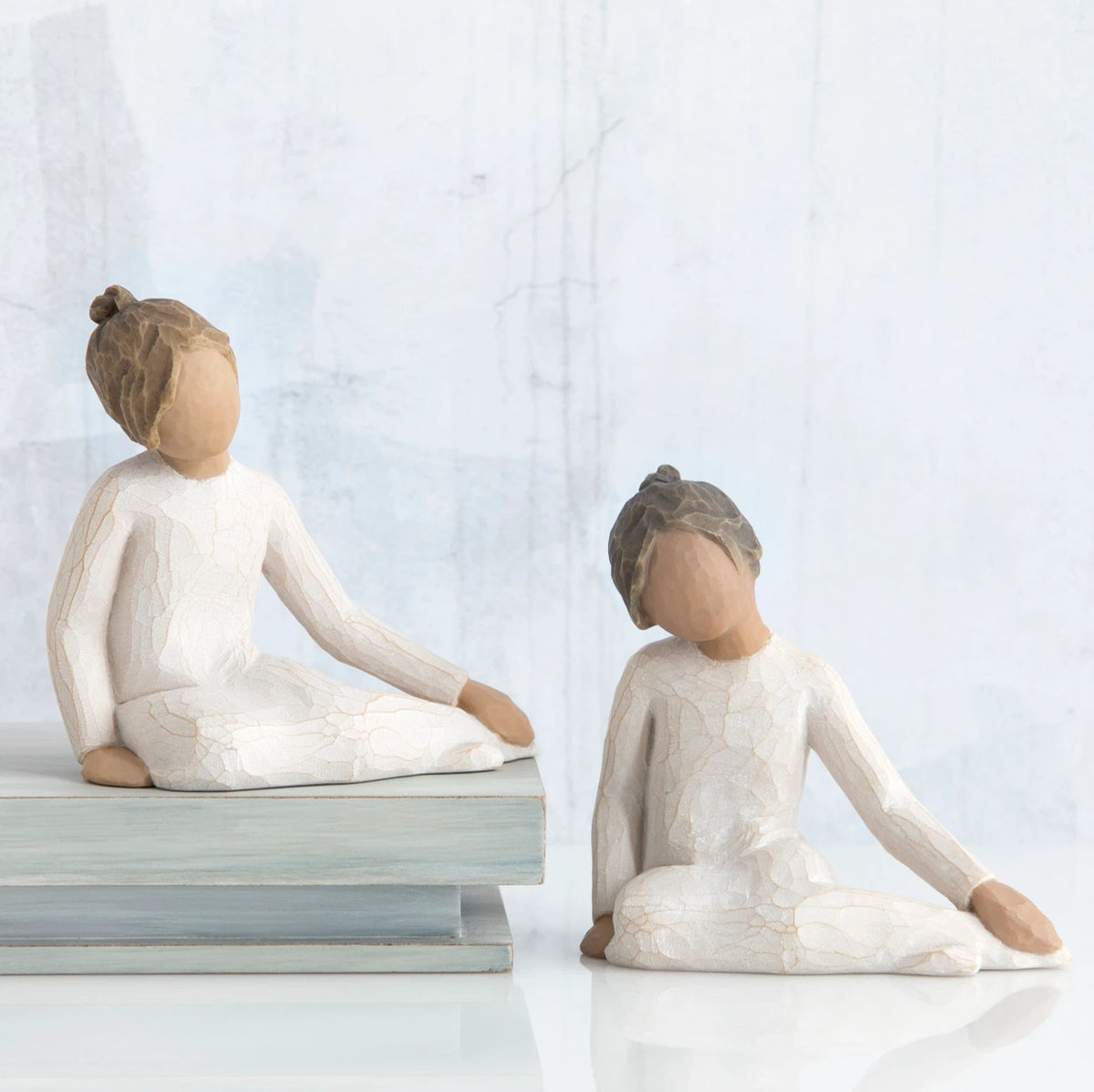 Two young brunette girl figurines in white sitting down next to wooden stack of books