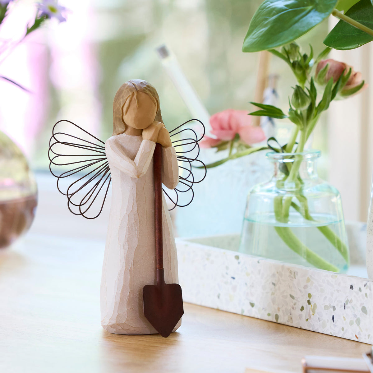 Willow Tree Angel of the Garden, 5.5”h hand-painted standing resin figure in cream dress with wire wings, leaning against brown metal shovel, on table with florals